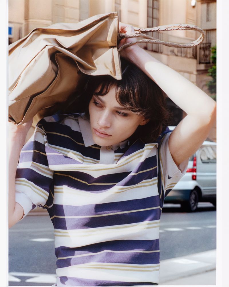 Model wearing a striped polo shirt holding a large beige paper bag overhead on a street