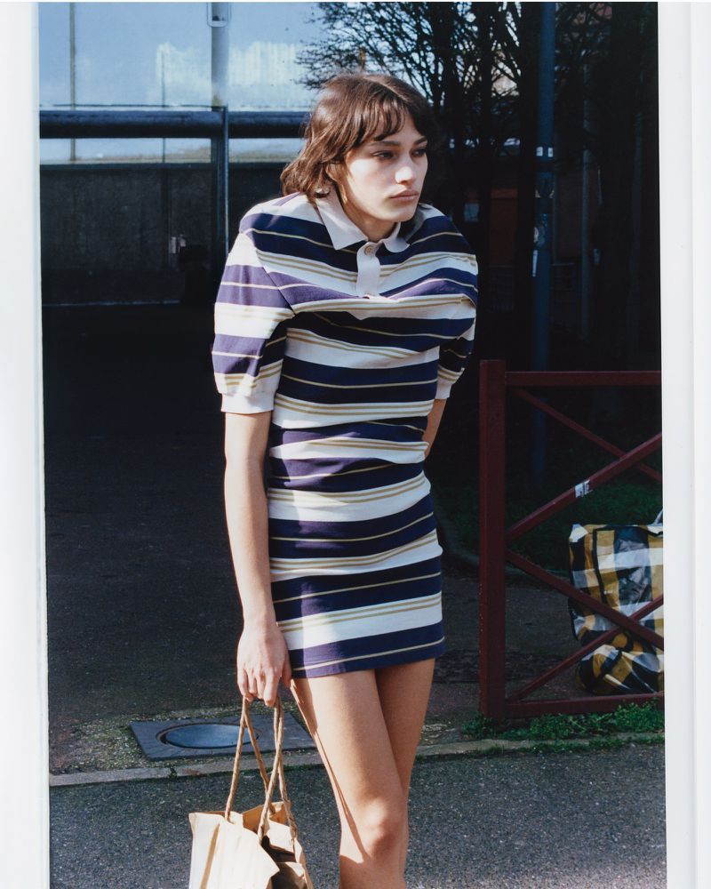 A woman in a striped polo mini dress holds a brown paper bag outdoors in an editorial photograph.