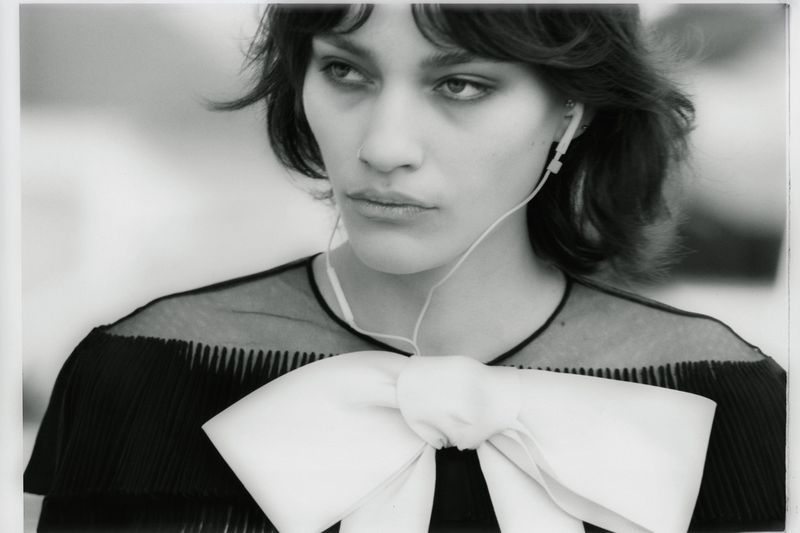 Black and white portrait of a woman wearing wired earphones and a black pleated top with an oversized white bow.