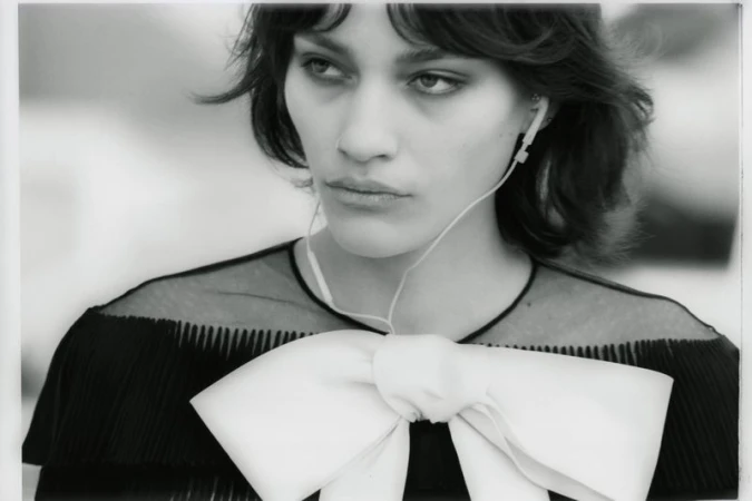 Black and white portrait of a woman wearing wired earphones and a black pleated top with an oversized white bow.