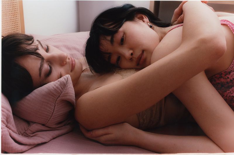 Two women lying together on a pink bed with arms embraced in an intimate domestic bedroom setting