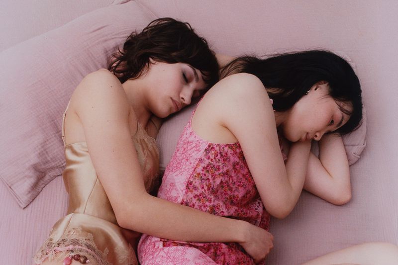 Two women asleep in close embrace on a pink bed wearing a champagne lace camisole and a pink floral slip dress.