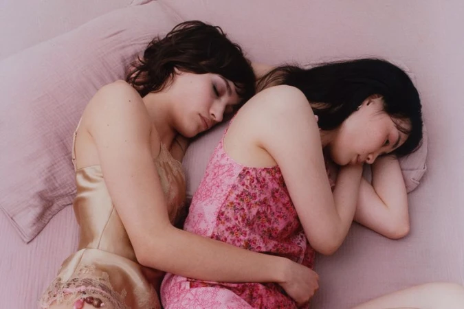 Two women asleep in close embrace on a pink bed wearing a champagne lace camisole and a pink floral slip dress.