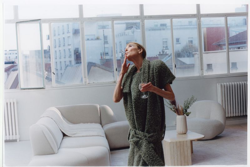 Elderly woman in green textured dress holding a glass while smoking inside a bright living room with large windows