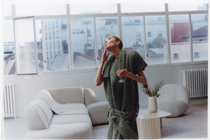 Elderly woman in green textured dress holding a glass while smoking inside a bright living room with large windows