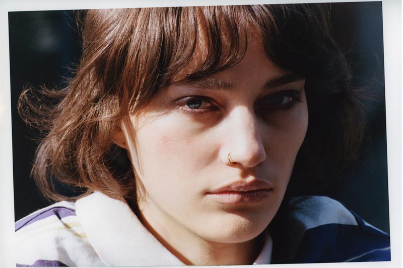 Close-up portrait of a woman with short brunette hair, bangs, and a gold nose ring wearing a striped collared shirt in direct sunlight.