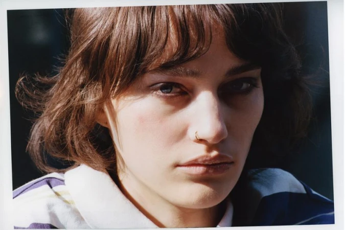 Close-up portrait of a woman with short brunette hair, bangs, and a gold nose ring wearing a striped collared shirt in direct sunlight.