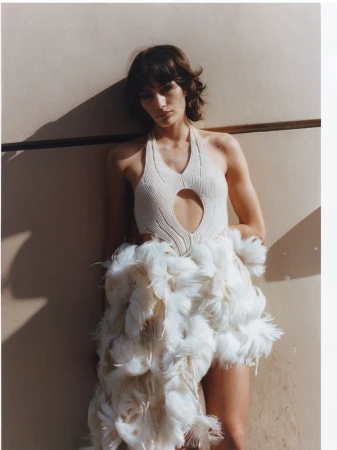 Model posing against a sunlit wall wearing an off-white cutout halter knit top and a white feathered skirt.