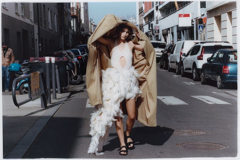 Model walking down a city street wearing a white feathered dress and lifting an oversized tan coat overhead.