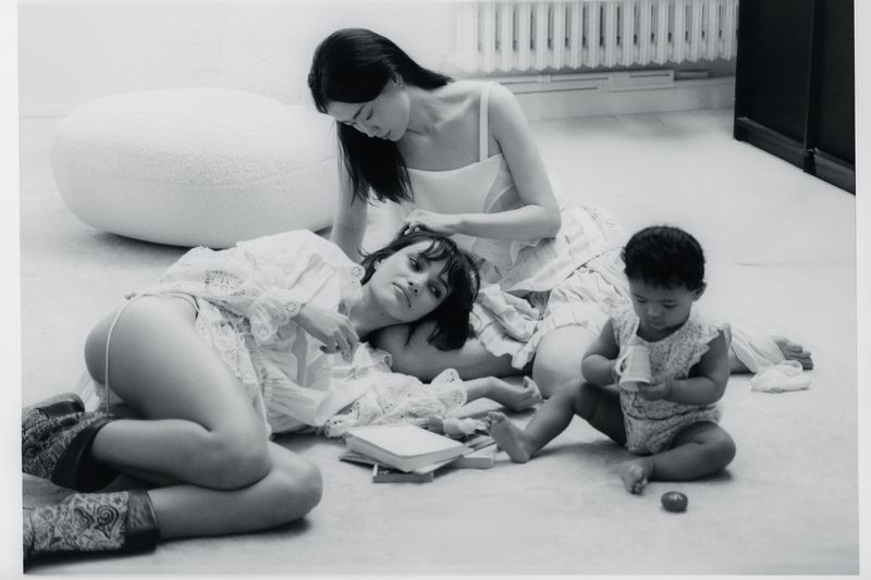 Black and white photo of two women and a toddler sitting on the floor with books and a white round ottoman in a bright room.