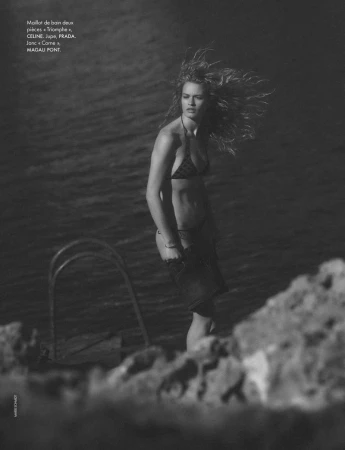 Model Britt Oosten posing in a bikini with windswept hair near a metal swim ladder by the water in black and white.