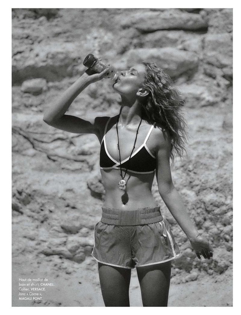 Black and white fashion portrait of Britt Oosten drinking from a bottle wearing a bikini top and running shorts against rocks.