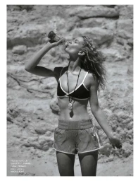 Black and white fashion portrait of Britt Oosten drinking from a bottle wearing a bikini top and running shorts against rocks.
