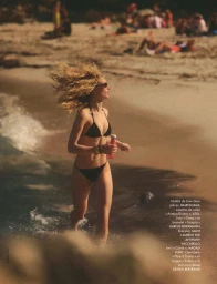 Britt Oosten running along a sandy beach shoreline wearing a black bikini and dark sunglasses while holding a pink bottle.