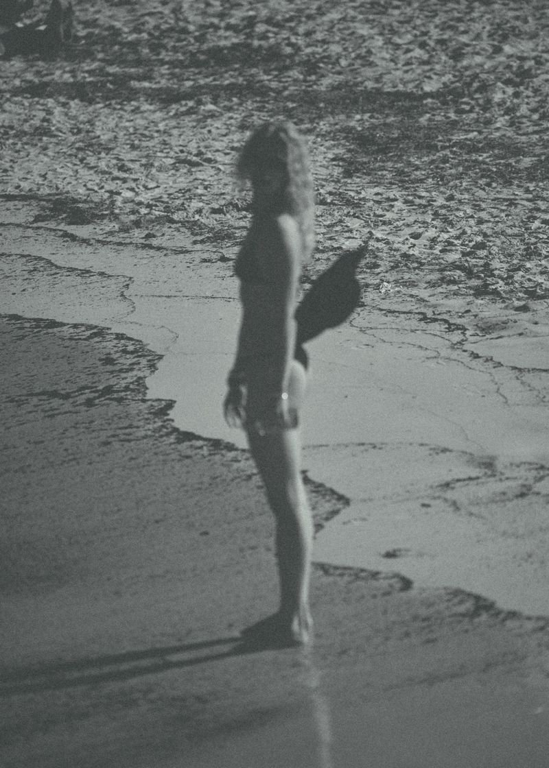 Monochrome grainy photograph of a woman in swimwear standing along the sandy shoreline holding a wide-brim hat.