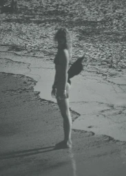 Monochrome grainy photograph of a woman in swimwear standing along the sandy shoreline holding a wide-brim hat.