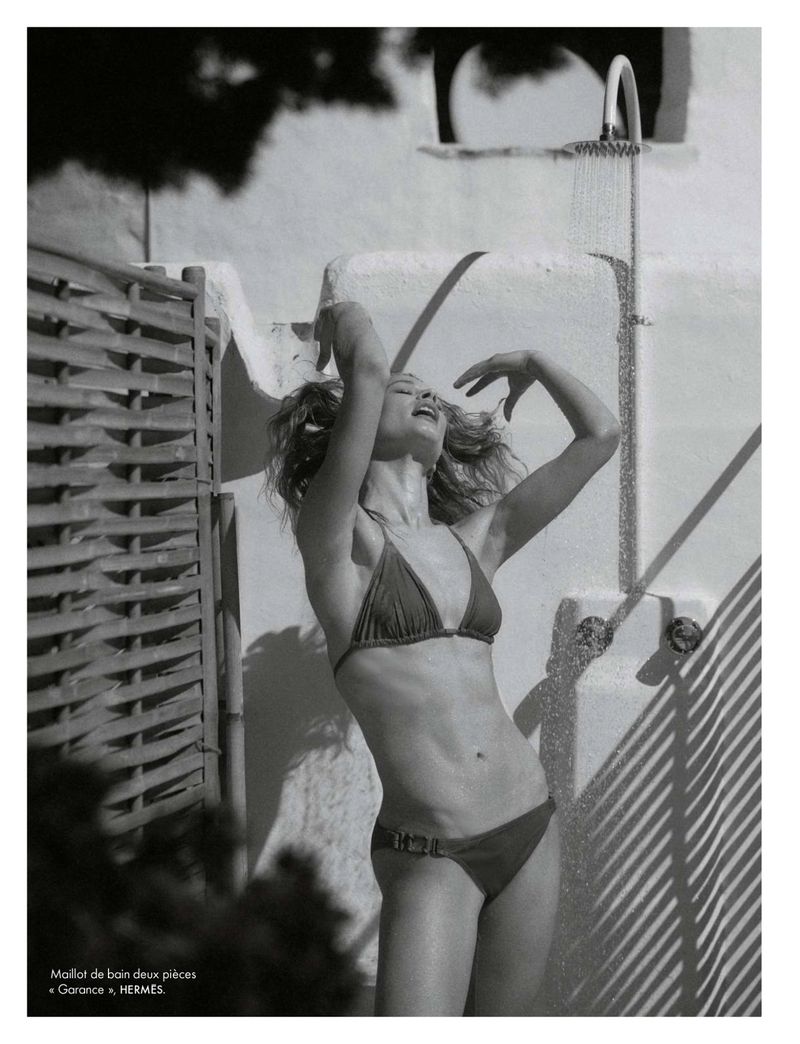 Model Britt Oosten wearing a two-piece bikini with hands raised while standing under an outdoor shower in direct sunlight.