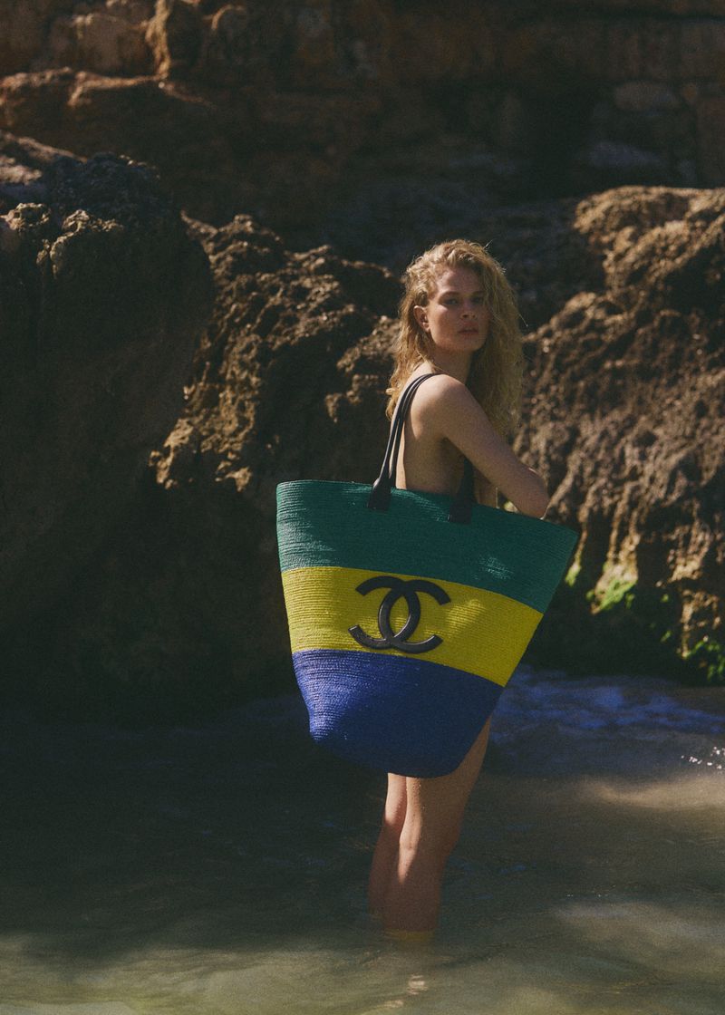 Model Britt Oosten standing in shallow coastal water carrying an oversized striped woven Chanel tote bag against rugged cliff rocks.