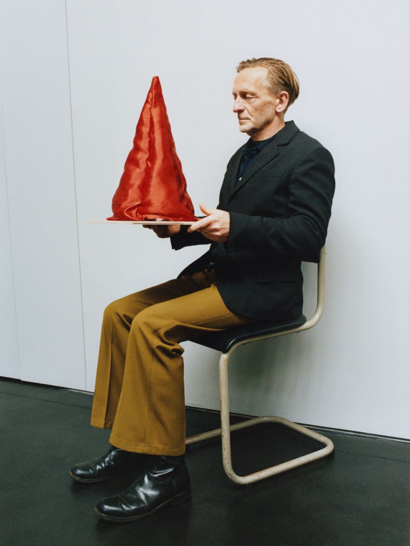Man in a dark blazer and mustard trousers seated on a chair holding a red conical fabric object against a white studio wall.