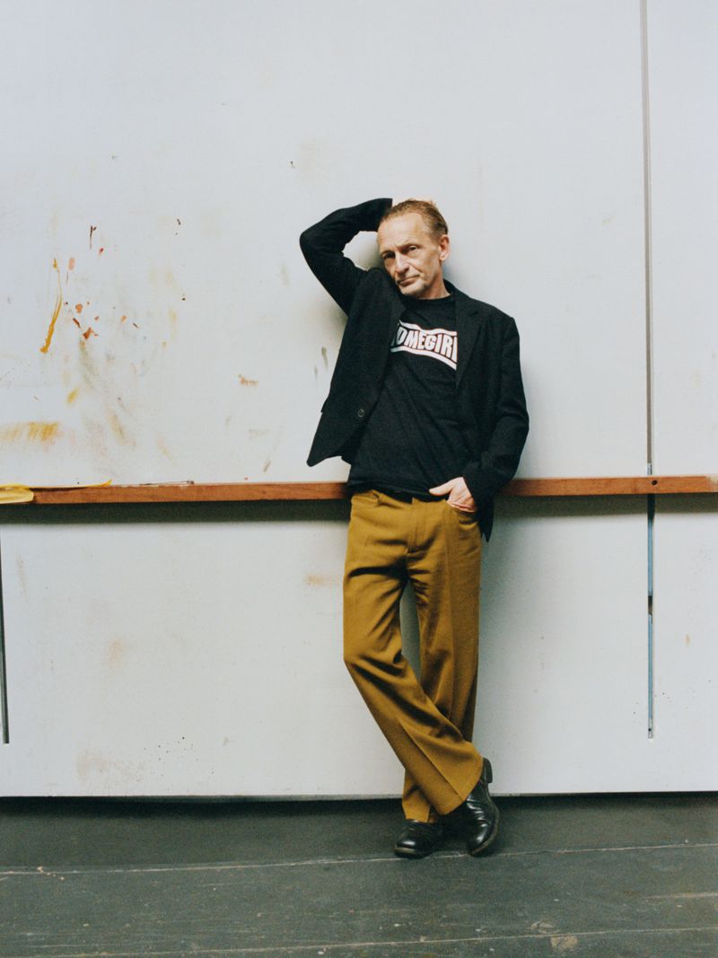 Man wearing black blazer, graphic t-shirt, and mustard trousers leaning against a white studio wall.