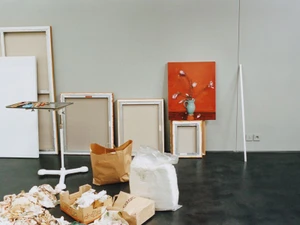 Artist studio space with stretched canvases, an orange floral still life painting, rolling palette stand, and supplies on a dark floor.