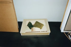 A small painted panel resting on a cardboard storage box on a dark studio floor flanked by art packaging.