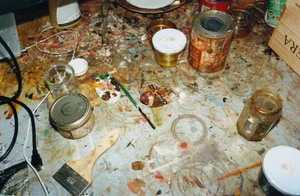Top-down view of an artist's paint-splattered workbench with paintbrushes, glass jars, tin cans, and dried pigments.
