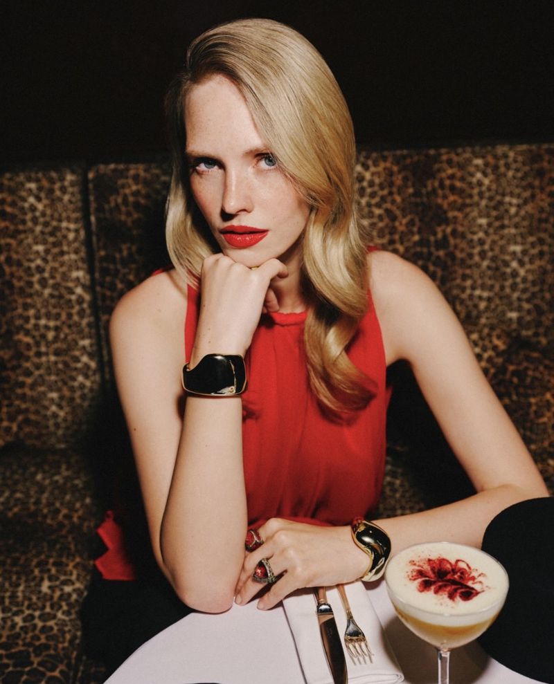 Model Charlotte Nolting in a red sleeveless top and gold cuff bracelets seated at a table with a cocktail in front of leopard print.