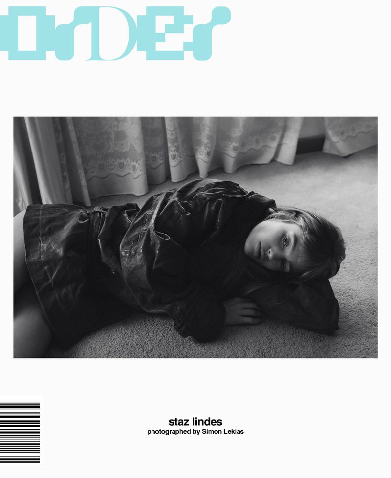 Black and white fashion magazine cover shot of a young woman lying on a carpet, with white curtains visible in the background.