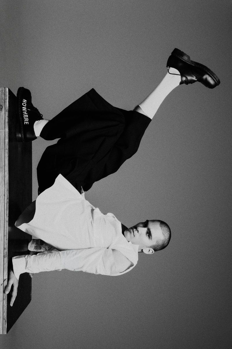 A man wearing a white shirt, black cropped trousers, and black shoes poses dynamically on a wooden platform against a studio background.