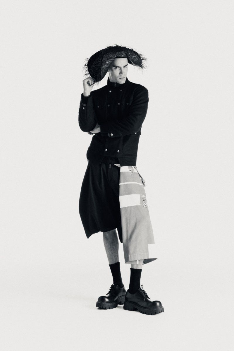 Fashion model wearing a black structured jacket, avant-garde layered skirt pants, and a wide textured hat against a white seamless backdrop.