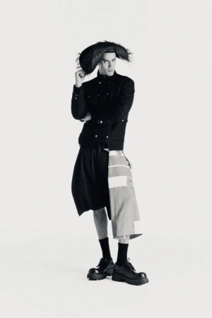 Fashion model wearing a black structured jacket, avant-garde layered skirt pants, and a wide textured hat against a white seamless backdrop.