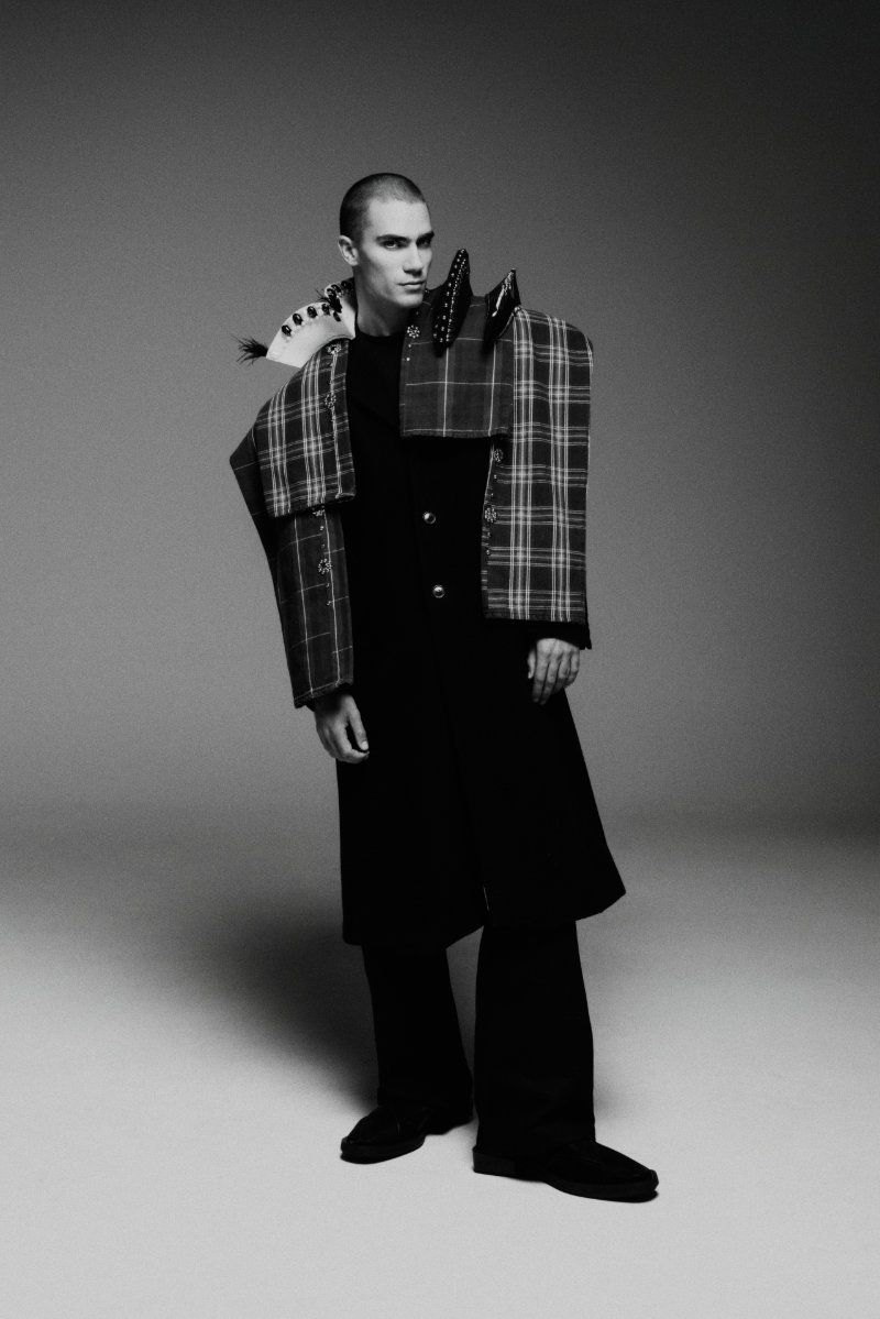 Man with buzz cut wearing an avant-garde deconstructed plaid and black coat against a neutral studio backdrop.