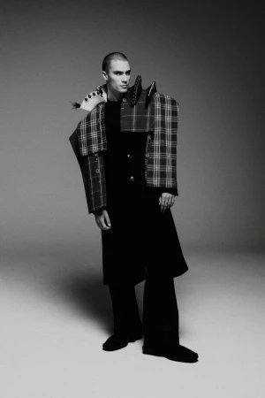 Man with buzz cut wearing an avant-garde deconstructed plaid and black coat against a neutral studio backdrop.