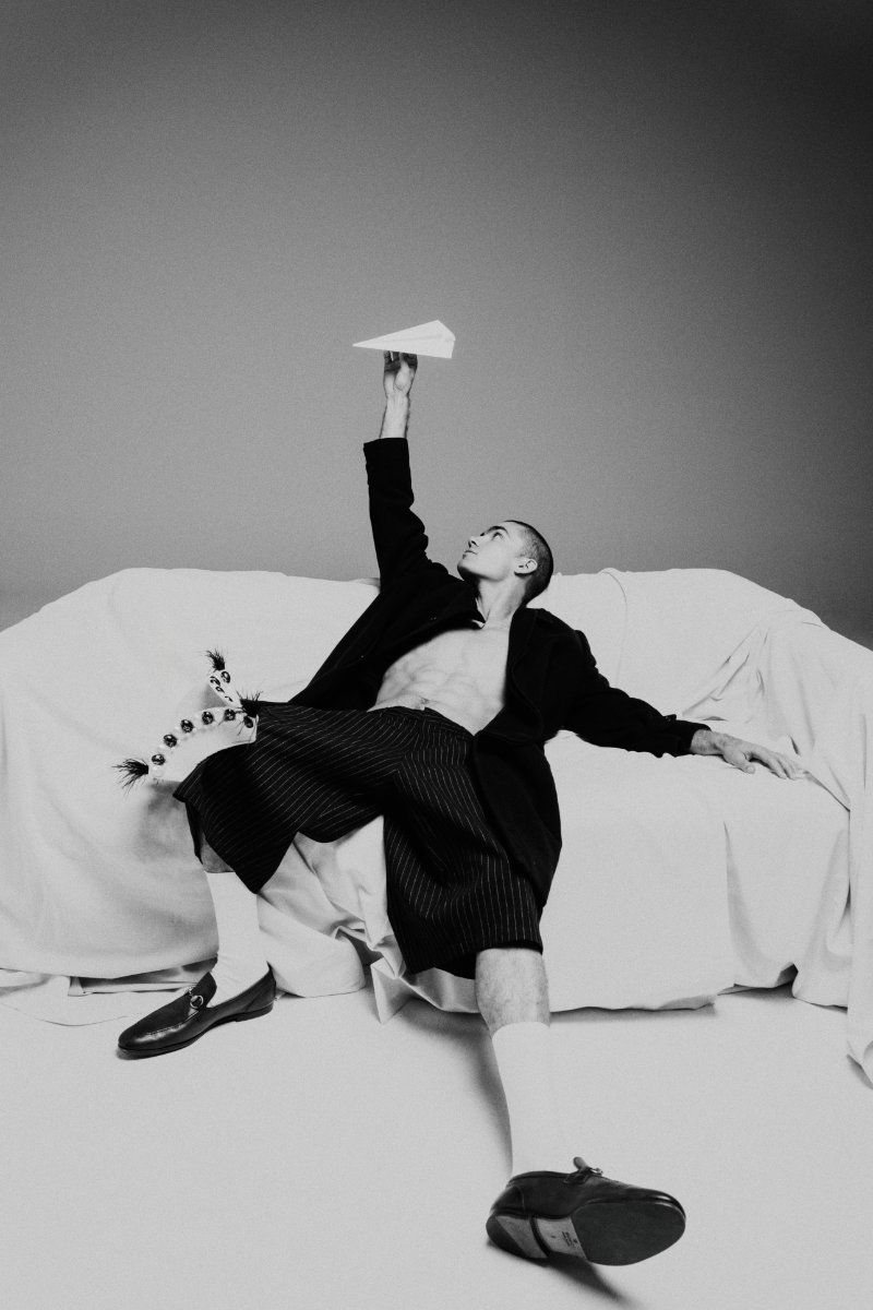 Buzz cut man in open dark coat and striped shorts reclines on a draped couch holding a paper airplane in a monochrome studio.