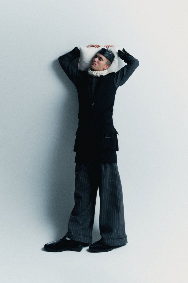 Model resting head against a white pillow wearing a black layered tunic and wide pinstriped trousers in a studio setting.