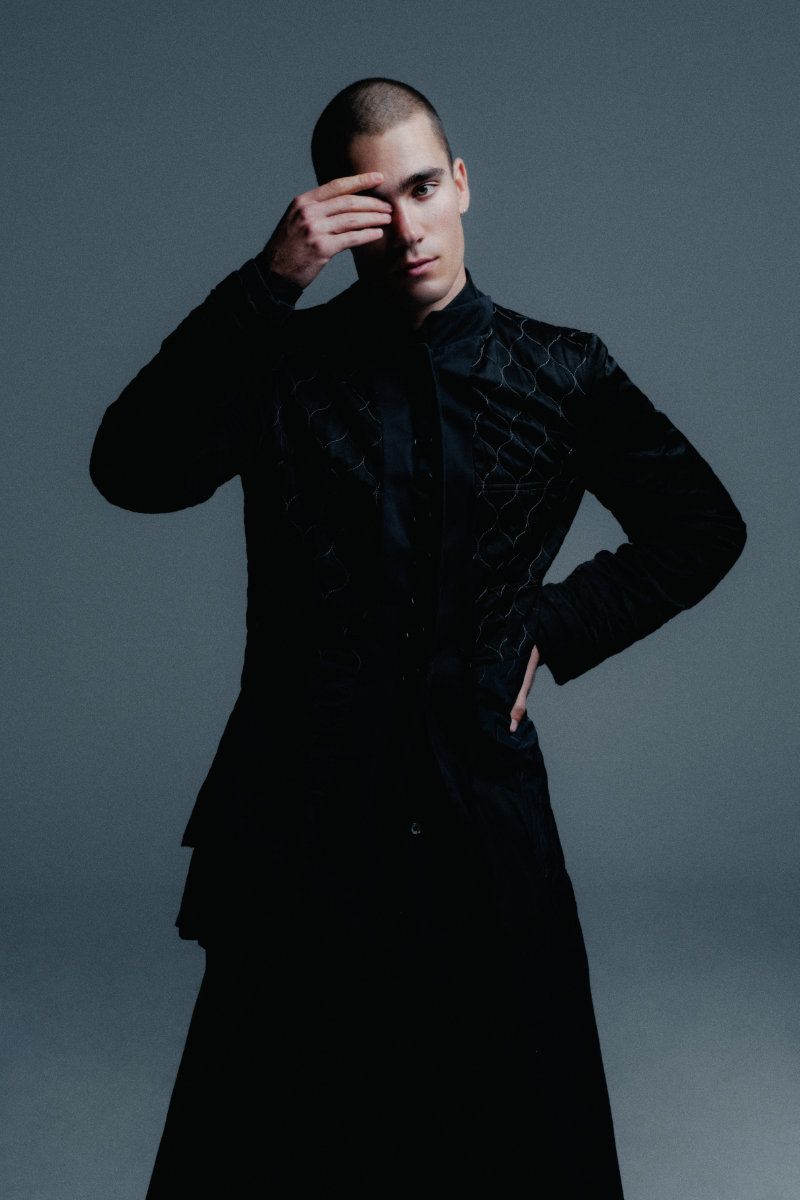 Male model with a buzz cut posing in a textured black long coat with one hand covering his eye against a grey studio backdrop.