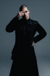 Male model with a buzz cut posing in a textured black long coat with one hand covering his eye against a grey studio backdrop.