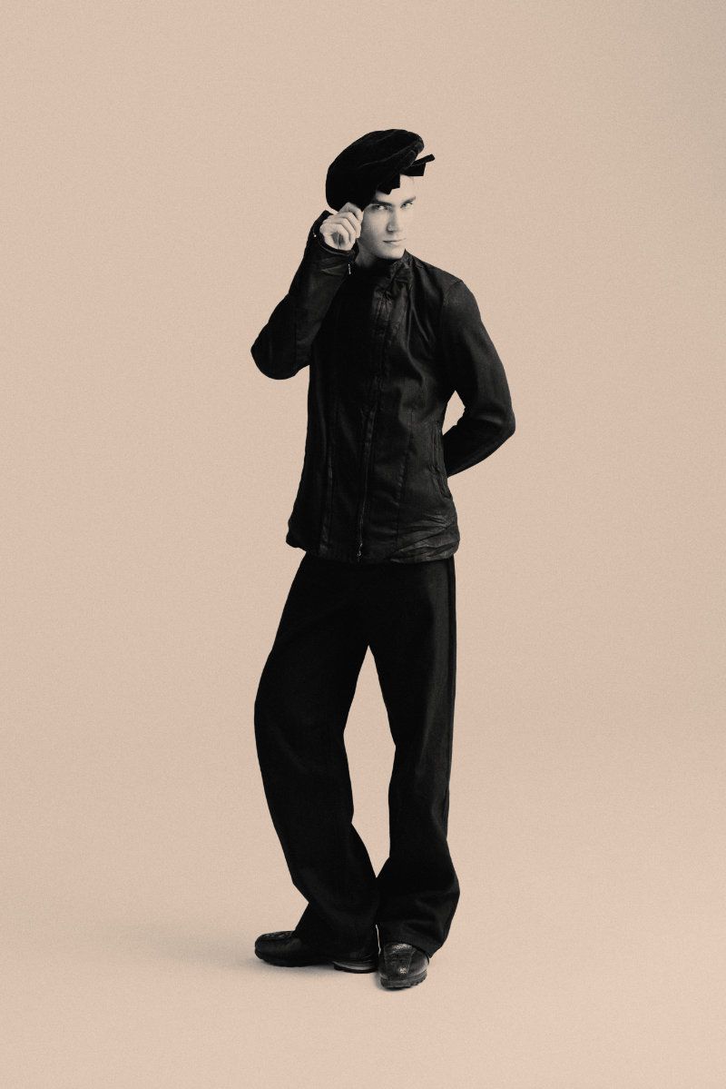 Full-length portrait of a man in a black jacket, loose trousers, and black beret standing against a neutral studio backdrop.