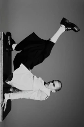 A man wearing a white shirt, black cropped trousers, and black shoes poses dynamically on a wooden platform against a studio background.
