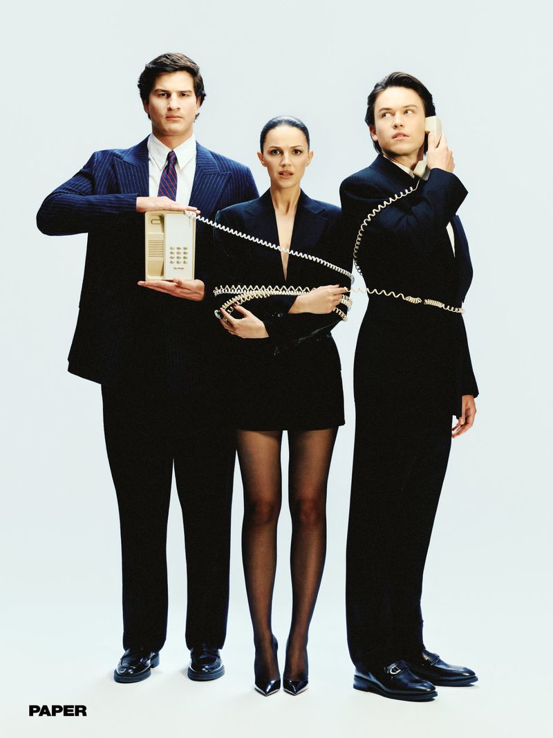 Three models dressed in black corporate suiting tangled together in a vintage telephone cord against a seamless white studio background.