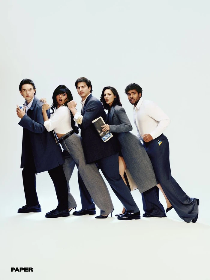 Five people in contemporary office attire posing in a diagonal leaning line against a plain white studio backdrop.