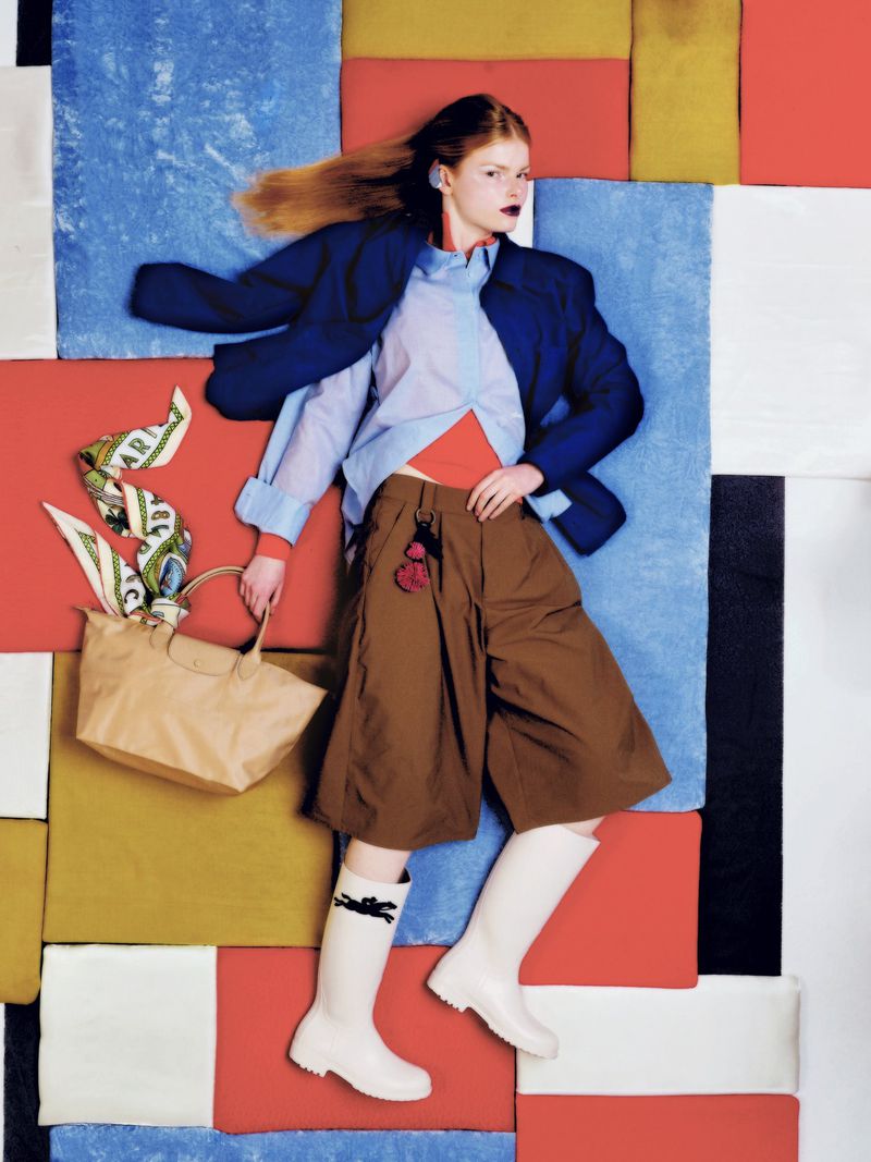 Model lying on a colorful geometric paneled floor wearing brown culottes, white boots, and holding a beige handbag