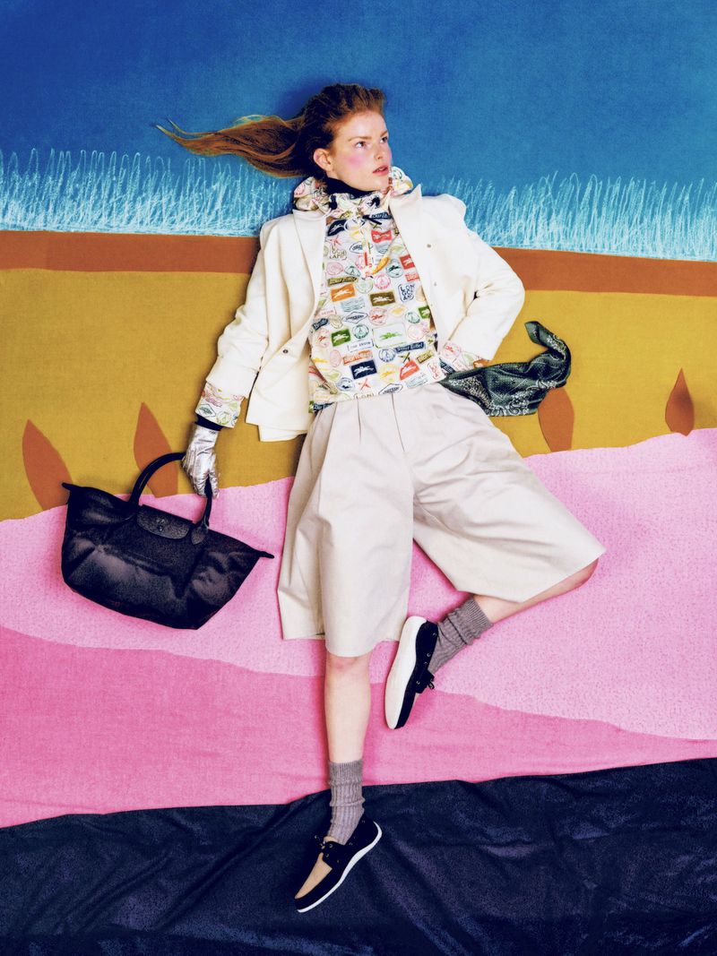 Model Elise Swain lying on a graphic colorblock fabric backdrop in a white blazer, patterned top, beige shorts, and holding a black tote.