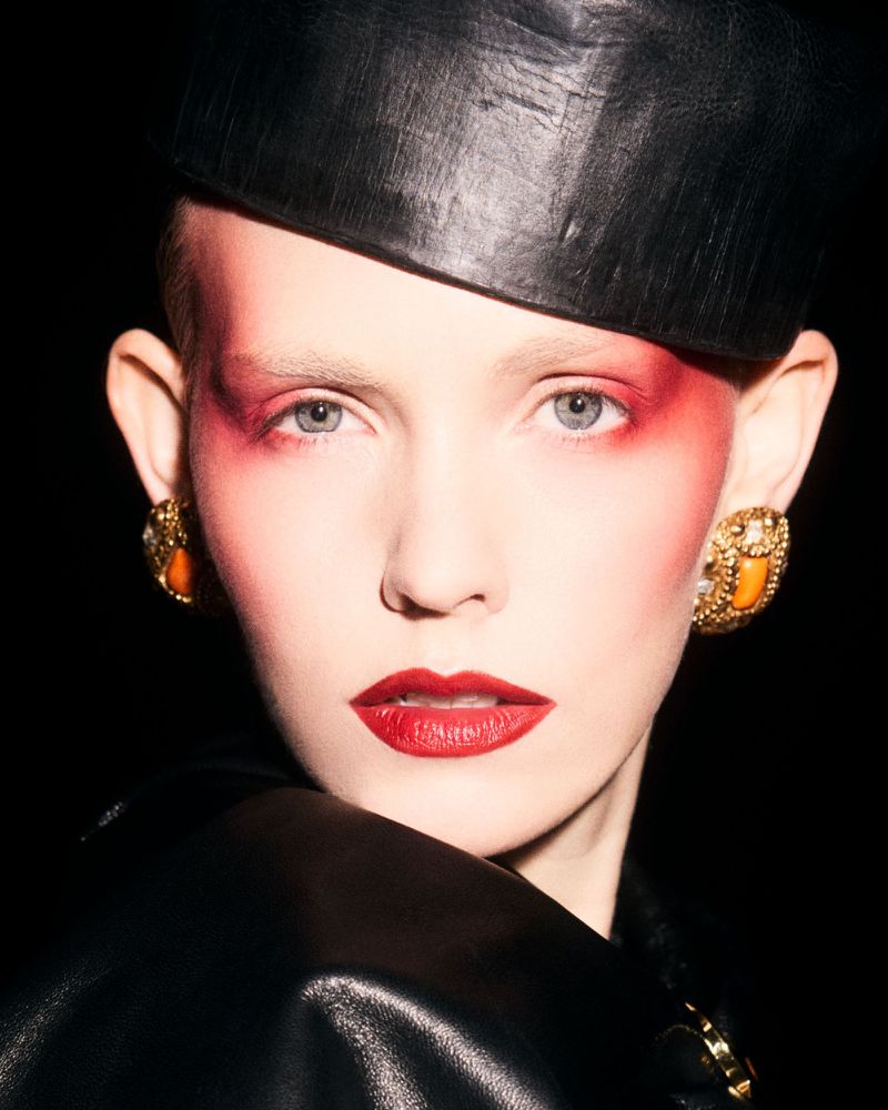 Close-up beauty portrait of Charlotte Nolting with bold red eyeshadow, red lipstick, gold statement earrings, and a black leather cap.