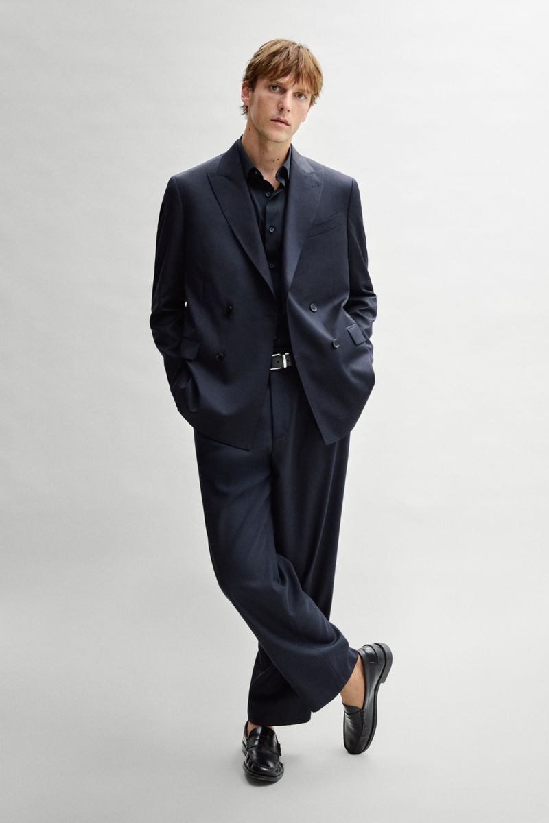 Model Quentin Demeester wearing an oversized navy double-breasted suit and black loafers against a light grey studio background.