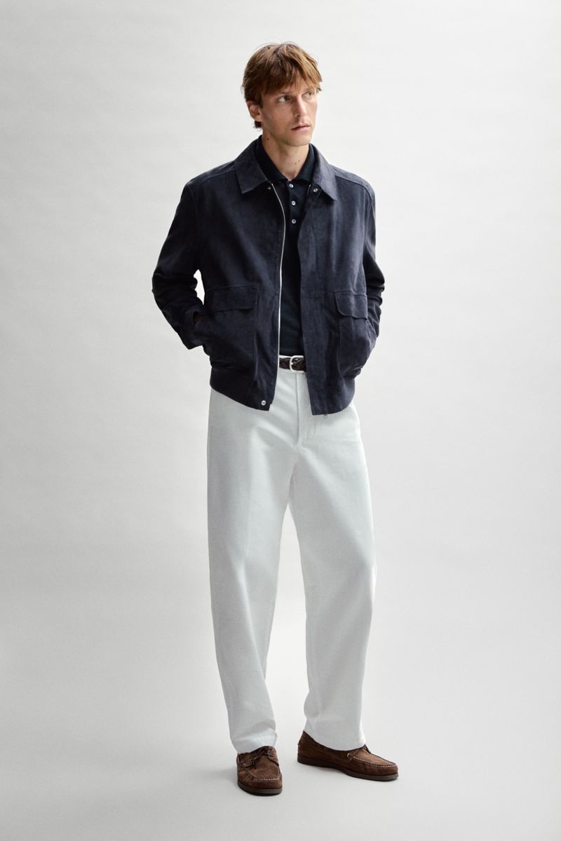 Quentin Demeester poses in a dark navy zip-up jacket, relaxed white trousers, and brown loafers against a pale studio backdrop.