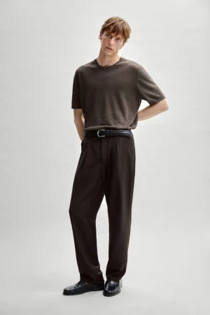 Model Quentin Demeester in a brown knit t-shirt, dark pleated trousers, and black loafers against a neutral grey studio backdrop.