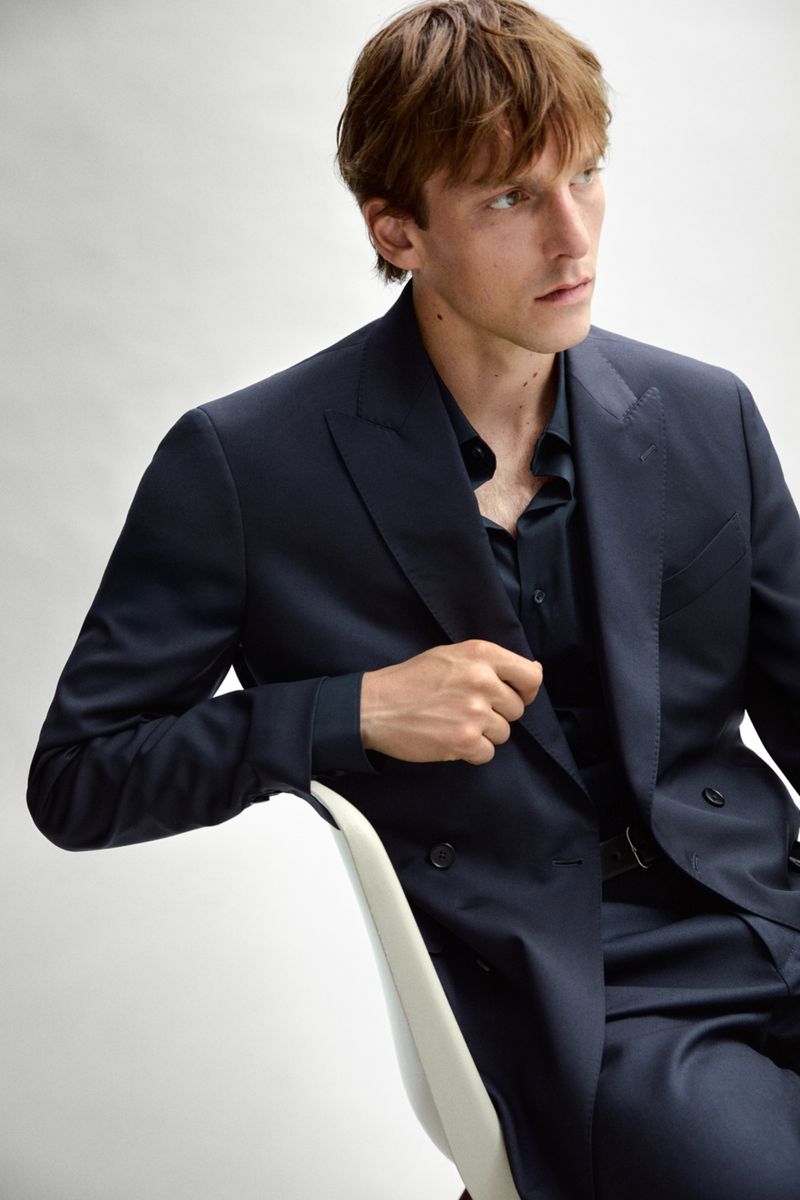 Model Quentin Demeester seated in a navy double-breasted suit against a clean white studio backdrop.