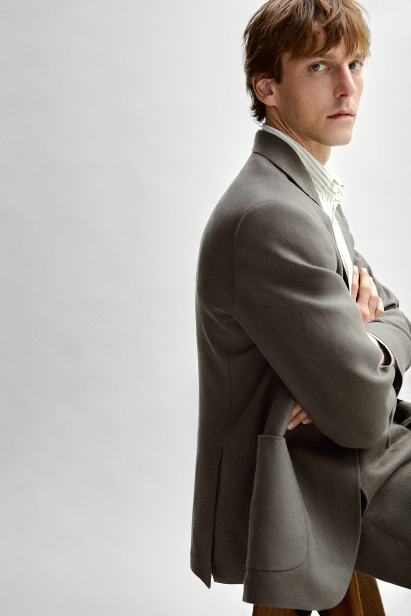Model Quentin Demeester wearing a tailored taupe blazer and striped collared shirt against a light grey seamless backdrop.